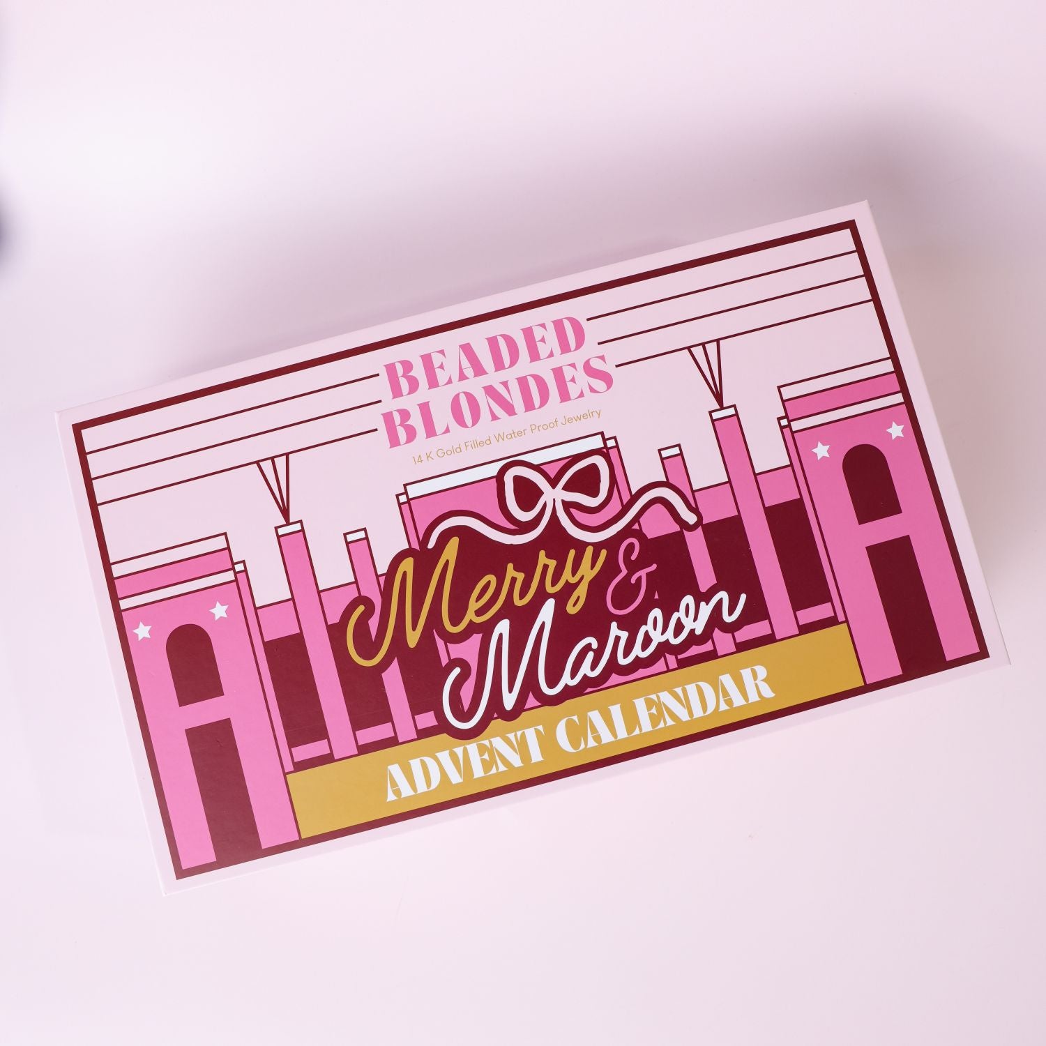 A rectangular advent calendar box titled 'Beaded Blondes Merry & Maroon Advent Calendar' shown at an angle on a light pink background. The box features a pink and burgundy football stadium illustration with gold and white typography.