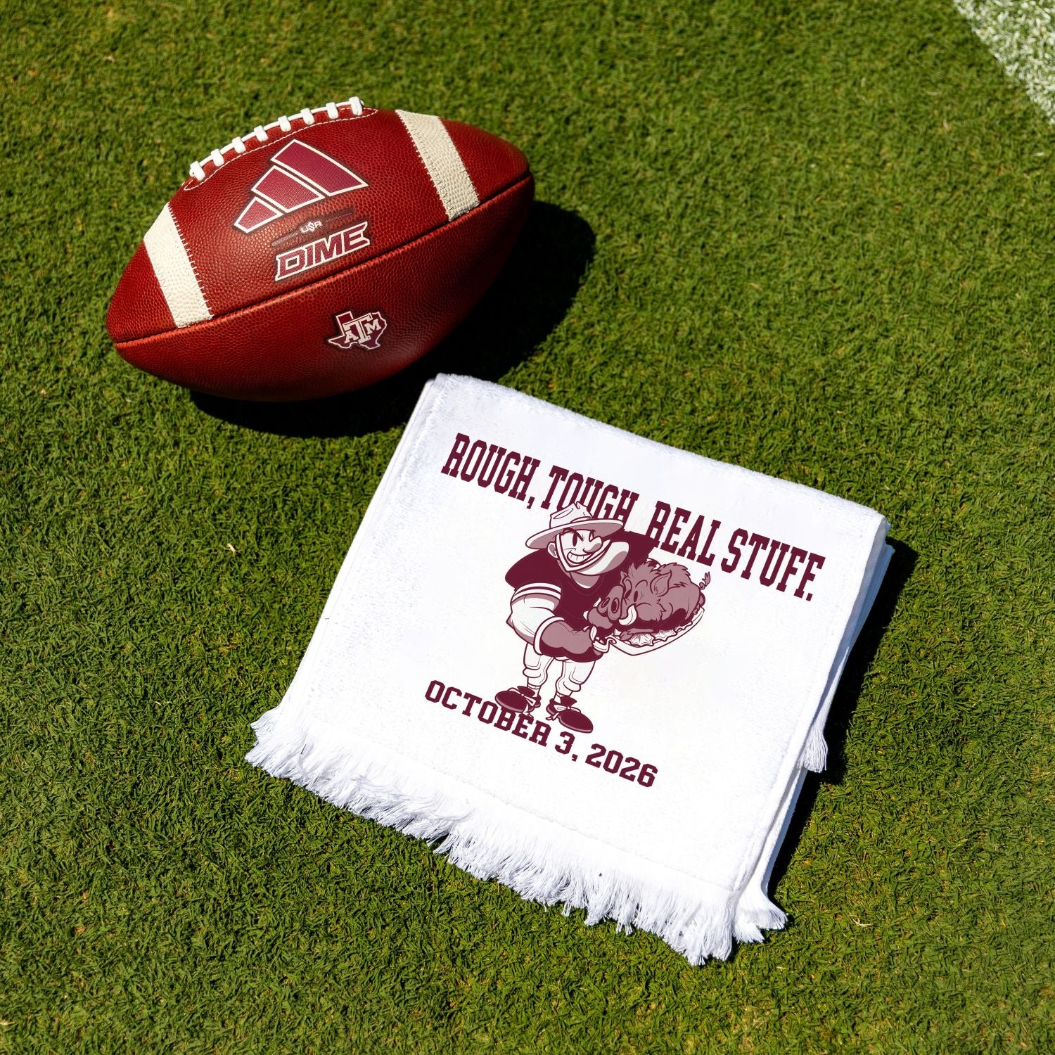 A white fringed rally towel with maroon text and a cartoon football player graphic lies on green grass next to a brown leather football.