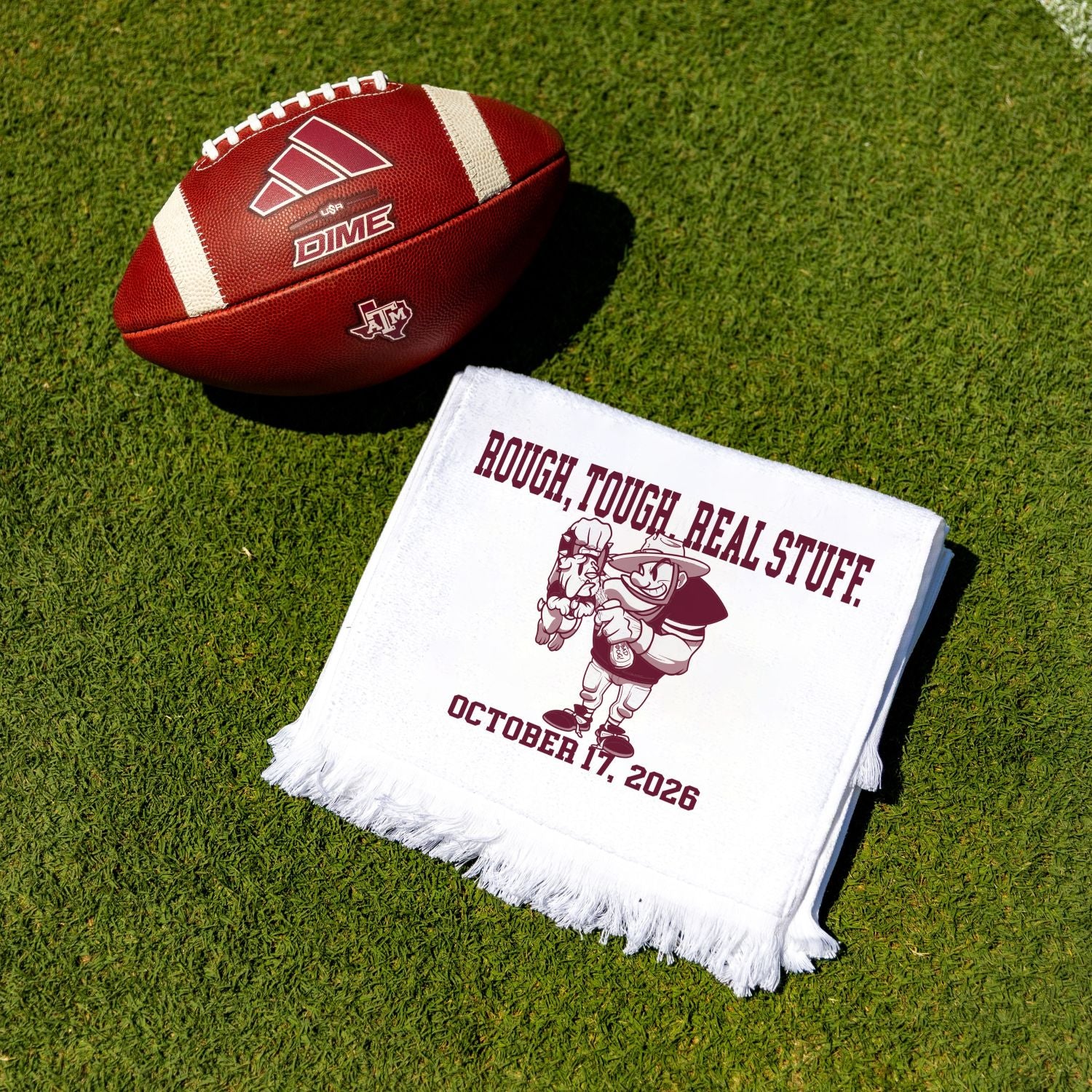 A white fringed towel with maroon printed text and mascot artwork lies on green turf beside a brown leather football. The blanket reads “ROUGH TOUGH REAL STUFF.