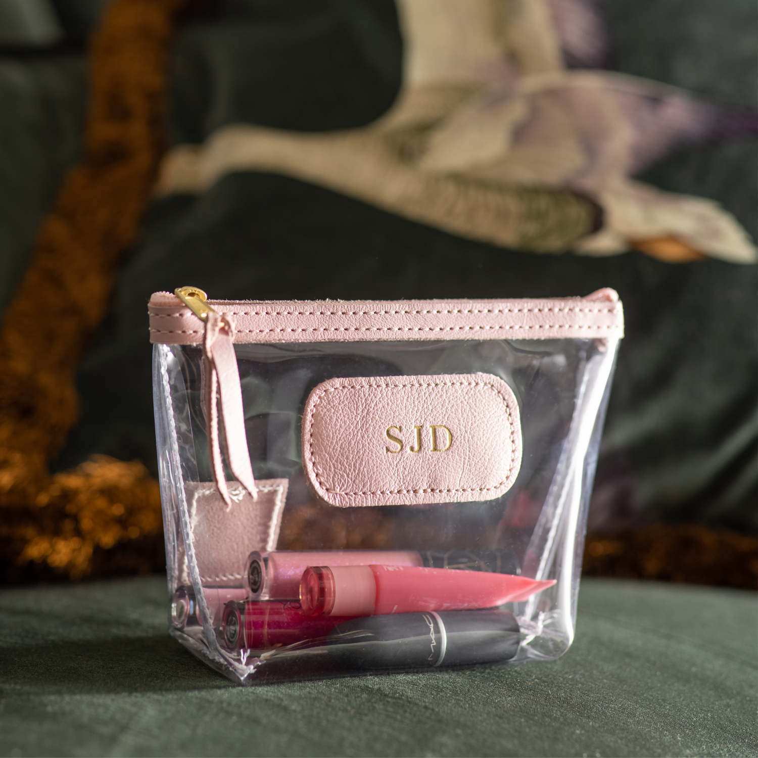 Clear makeup bag with pink zipper and monogrammed patch containing 'SJD' on a green surface.