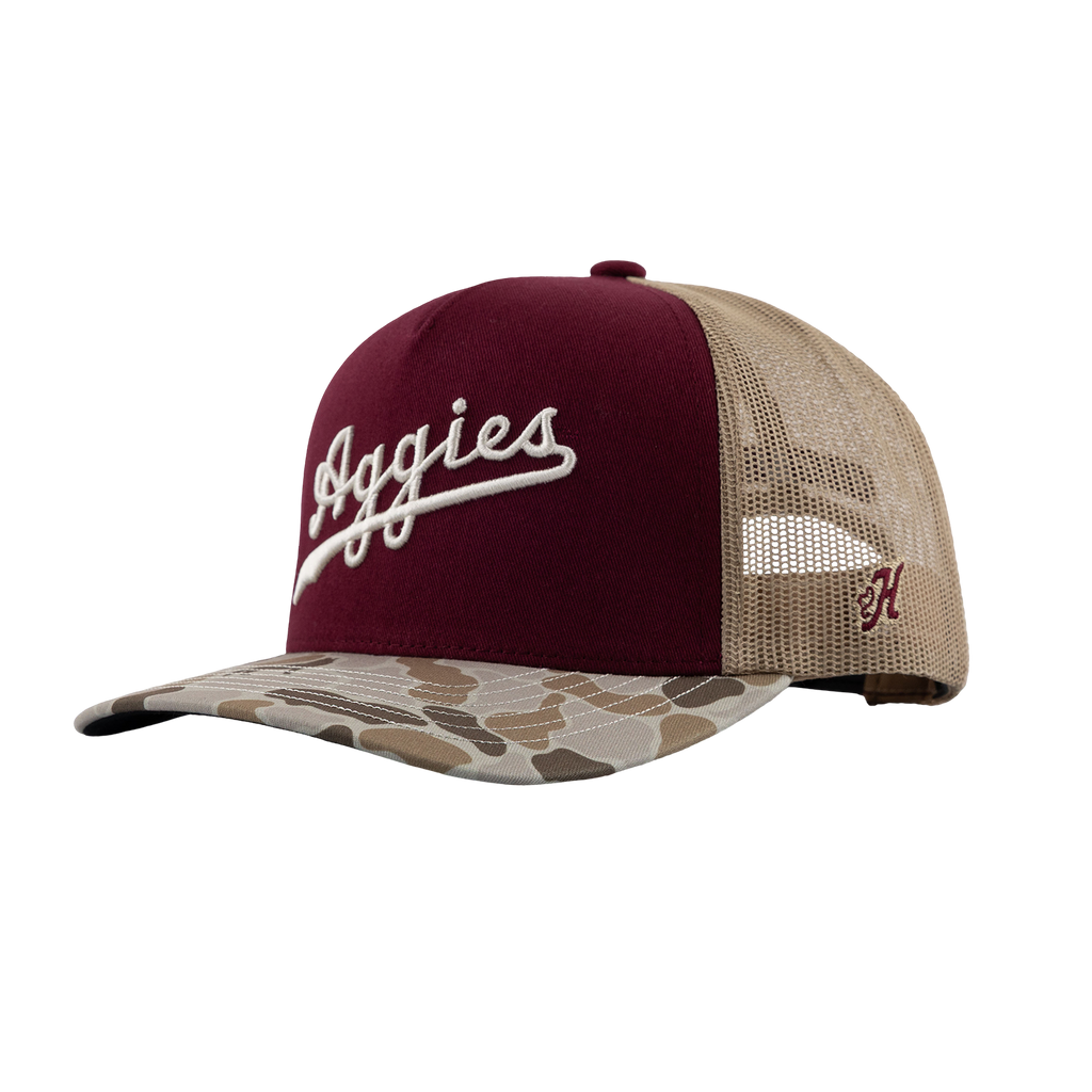 A trucker-style baseball cap with a maroon front panel, beige mesh back, and a camouflage-patterned brim. The front features white embroidered script reading 'Aggies', and the side displays a small maroon embroidered Hooey brand 'H'.