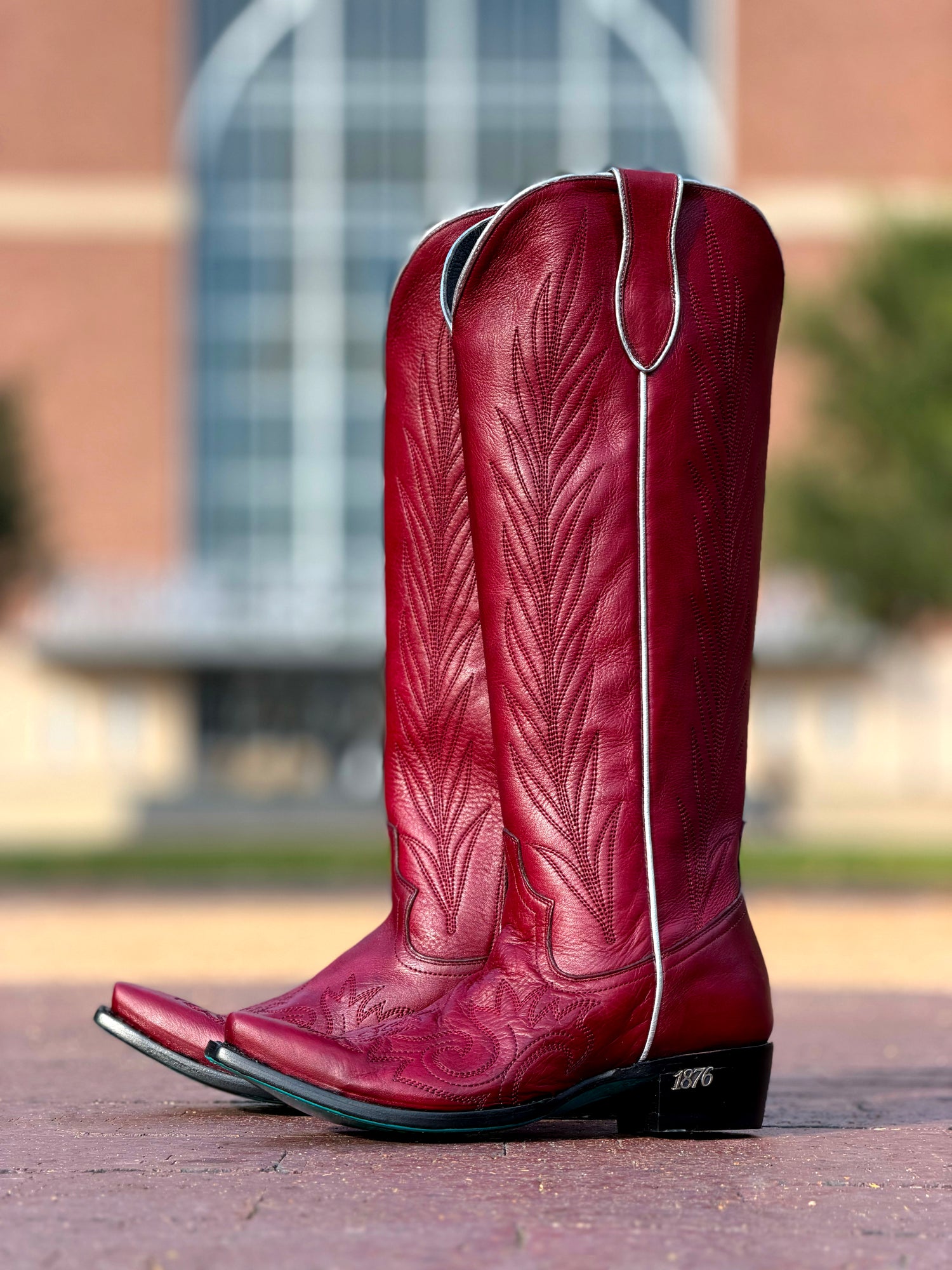 Maroon Texas A&M-inspired cowboy boots featuring Western-style embroidered detailing, silver piping, pointed toes, and black heels marked “1876.”