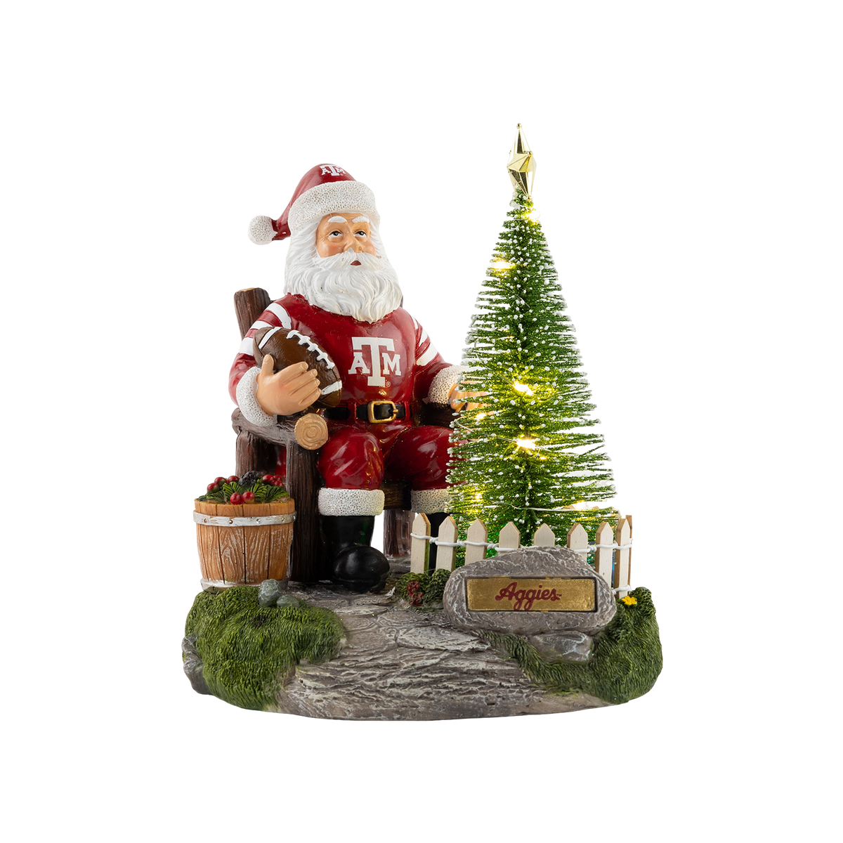 A Christmas figurine of Santa Claus wearing a maroon Texas A&M suit and holding a football, seated next to an illuminated Christmas tree behind a white picket fence with an "Aggies" sign.   