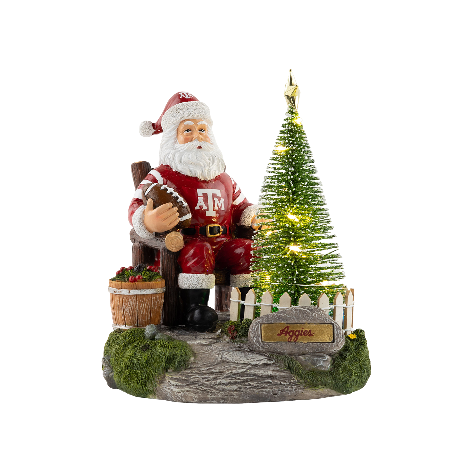 A Christmas figurine of Santa Claus wearing a maroon Texas A&M suit and holding a football, seated next to an illuminated Christmas tree behind a white picket fence with an "Aggies" sign.   