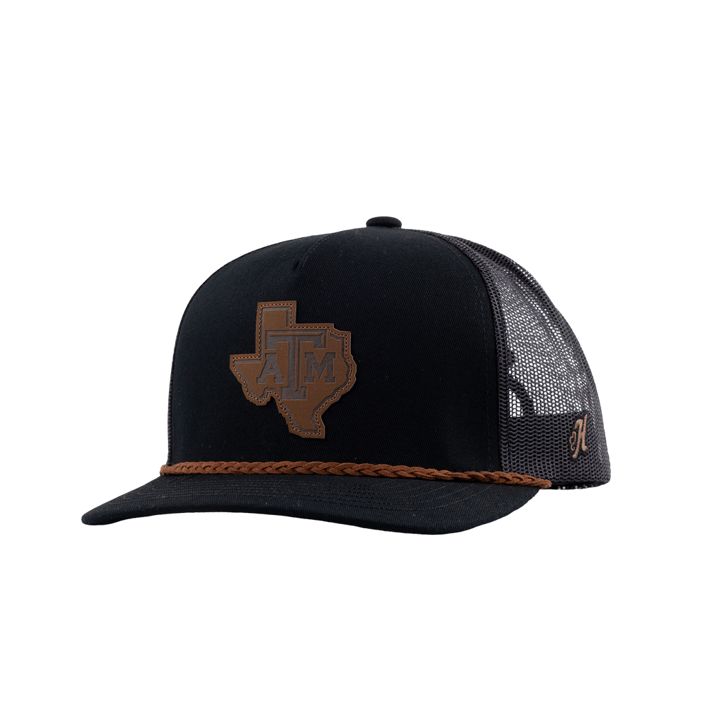 A black trucker hat with a brown leather Texas A&M patch on the front, a braided brown leather cord detail along the brim, and a brown mesh back with Gig 'Em embroidery.