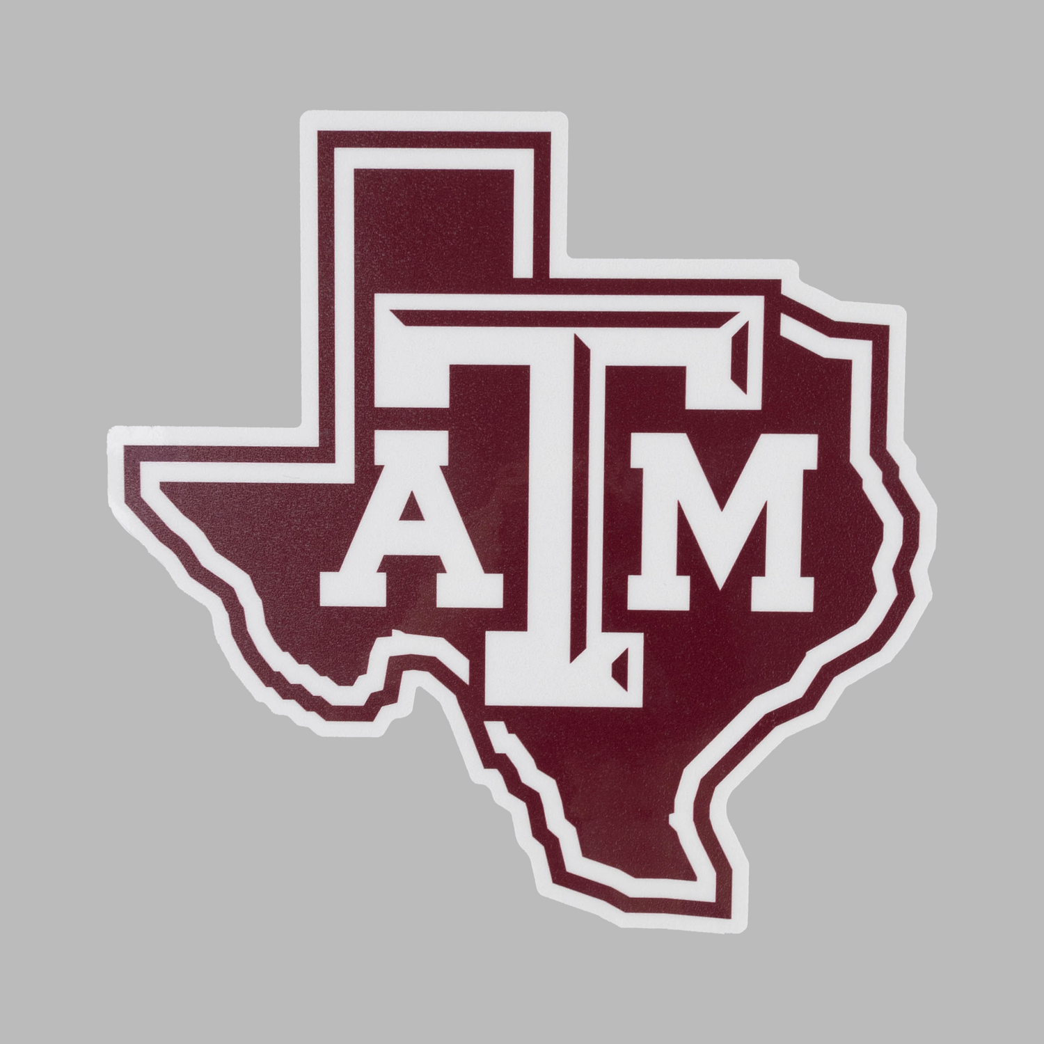 A die-cut sticker shaped like the state of Texas, featuring the letters 'A' and 'M' in white over a dark maroon background, with a white border, shown on a solid gray background.