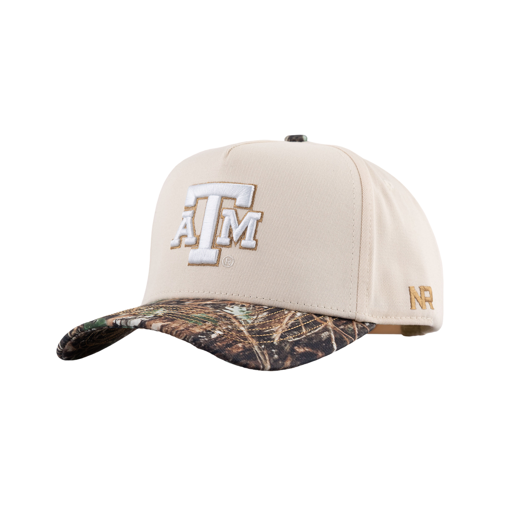 A baseball cap featuring a cream-colored crown with a large white and gold 'A&M' logo on the front, and a camouflage-patterned visor. A small gold 'NR' logo is visible on the side.